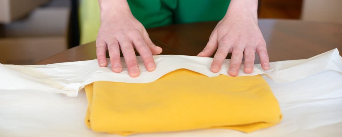 Mãos embalando uma peça de roupa amarela em papel, sobre a mesa de uma loja online